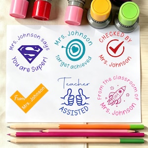 May include: Six circular self-inking stamps with colorful caps, displaying various teacher-themed designs and text including Mrs. Johnson says You are Super, target achieved, CHECKED BY, From, Teacher ASSISTED, and From the classroom of Mrs. Johnson.