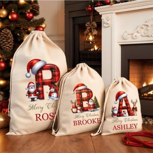 Pode incluir: Três sacolas de presente de Natal bege com fechos de cordão. Cada sacola apresenta uma letra grande em um padrão xadrez vermelho e preto, encimada por um gorro de Papai Noel. As sacolas são decoradas com o Papai Noel, um boneco de neve e o texto "Merry Christmas" e um nome.