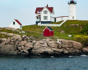 May include: A coastal scene featuring a white lighthouse with a black top, a white house with a red roof, and a small red building. The structures sit on a grassy hill above rocky terrain, with the ocean in the foreground and a cloudy sky.