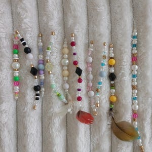 Può includere: Una collezione di ornamenti di perline con vari colori e forme. Gli ornamenti presentano perle, perline colorate e dettagli dorati. Alcuni hanno piccole piume attaccate. Gli ornamenti sono esposti su una superficie bianca strutturata.