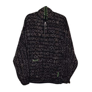 Op de afbeelding: Zwarte jas met ritssluiting en een herhalend patroon van bruine cirkels en lijnen. De jas heeft een hoge kraag met neon groene stiksels en accenten. De rits is zilverkleurig en er zijn neon groene kruissteekdetails.