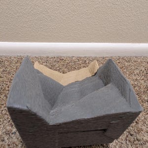 May include: A gray and brown cardboard box with a textured surface. The box is folded in half and has a rectangular shape.