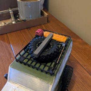 May include: A miniature green and white model SUV with a black roof rack. The rack holds two spare tires, a gray rectangular object, and orange and red accessories. The tires are black with a detailed tread.