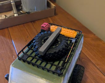 May include: A miniature green and white model SUV with a black roof rack. The rack holds two spare tires, a gray rectangular object, and orange and red accessories. The tires are black with a detailed tread.