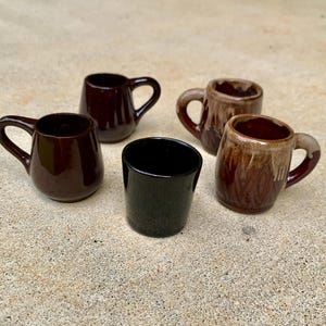May include: A collection of five small ceramic mugs in various finishes. Two mugs are solid dark brown, one is solid black, and two feature a two-tone brown glaze with a lighter, dripped effect near the rim and handle. Each mug has a single handle.