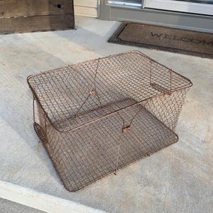 May include: A rectangular, vintage wire mesh basket with a copper-colored finish. The basket has a hinged lid and a handle on one side. The basket is sitting on a concrete surface.