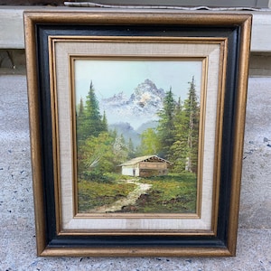May include: Oil painting of a mountain landscape with a small cabin, framed in gold and black. The artwork depicts a path leading to a cabin nestled among green trees, with a snow-capped mountain in the background. The frame is approximately 12 inches by 10 inches.