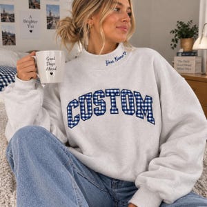 Puede incluir: Una sudadera gris claro de cuello redondo con la palabra CUSTOM en un patrón de cuadros vichy azul y blanco en el pecho. La zona del cuello incluye el texto bordado Your Name con un pequeño corazón. Una taza blanca con el texto Good Days Ahead.