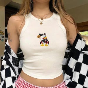以下が含まれることがあります： 黒いマントとシルクハットを身につけ、オレンジ色のジャック・オー・ランタンの風船を持ち、その上を小さなコウモリが飛んでいる、吸血鬼の格好をした漫画のキャラクターのアヒルが描かれた、クリーム色のノースリーブのタンクトップ。