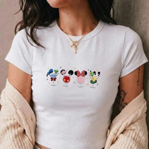 May include: A white short-sleeved t-shirt featuring a graphic print of four cocktail glasses, each stylized with iconic mouse ears and accessories, including a blue hat, a red bow, and a pink polka-dot bow, accented by small yellow star shapes.