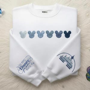 May include: White sweatshirt with embroidered Mickey Mouse heads in shades of blue across the chest. The left sleeve features the text "in my Disney mom era" and the right sleeve has a castle design, all in blue.