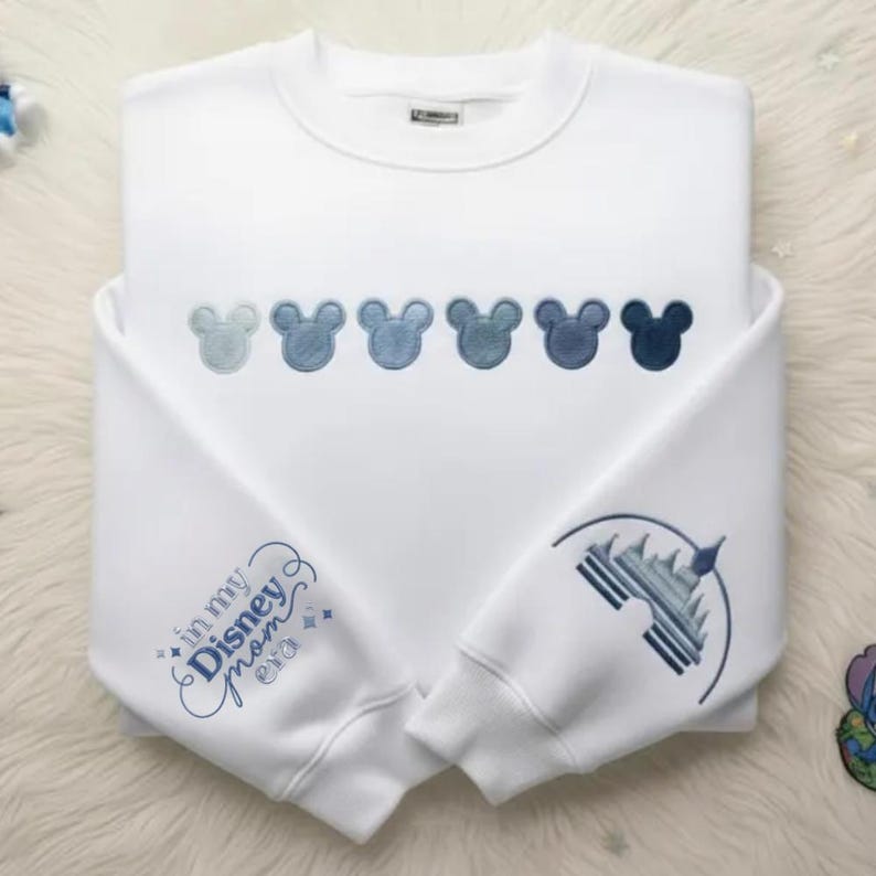 May include: White sweatshirt with embroidered Mickey Mouse heads in shades of blue across the chest. The left sleeve features the text "in my Disney mom era" and the right sleeve has a castle design, all in blue.