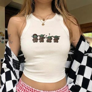 May include: White tank top with a graphic of four Grogu characters in brown robes. The first Grogu wears headphones, the second is gesturing, the third has a question mark, and the fourth is smiling.