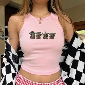 May include: A light pink ribbed tank top featuring a graphic of four Grogu characters in various poses. The characters are wearing brown robes. The tank top has a round neckline and a racerback design.
