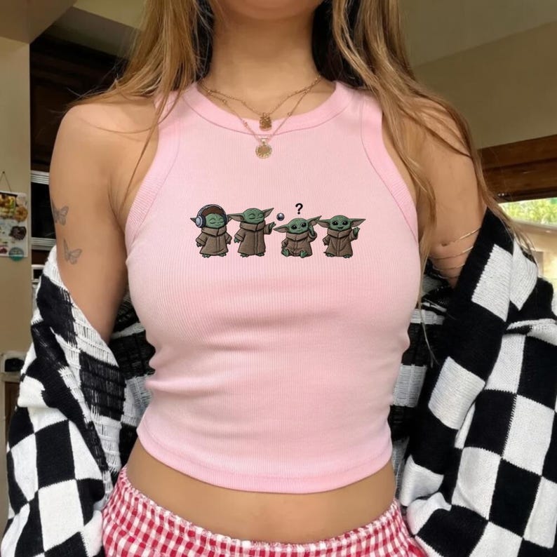 May include: A light pink ribbed tank top featuring a graphic of four Grogu characters in various poses. The characters are wearing brown robes. The tank top has a round neckline and a racerback design.