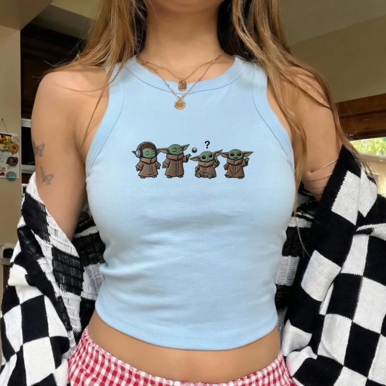 May include: Light blue tank top featuring a graphic of four Grogu characters in different poses. The Grogu characters are wearing brown robes. One Grogu has headphones, another is holding a ball, and one has a question mark above its head.