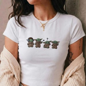 May include: White t-shirt featuring a cartoon graphic of four Grogu characters in various poses. One Grogu wears headphones, another has a ball, one has a question mark, and the last one is gesturing. The characters are in brown robes.