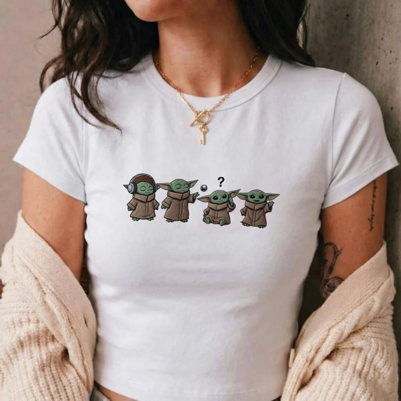 May include: White t-shirt featuring a cartoon graphic of four Grogu characters in various poses. One Grogu wears headphones, another has a ball, one has a question mark, and the last one is gesturing. The characters are in brown robes.