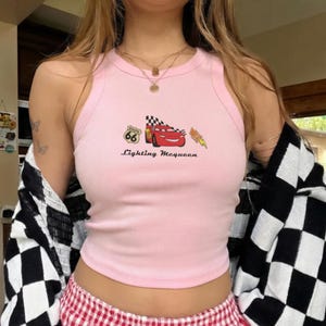 May include: A light pink ribbed tank top featuring a graphic of Lightning McQueen from the movie Cars. The graphic includes the Route 66 logo, a checkered flag, and the text "Lightning McQueen".