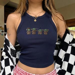 May include: Navy blue ribbed tank top featuring an embroidered design of four Grogu characters. The characters are in various poses, with one wearing headphones and another with a question mark above its head. The top has a round neckline and a sleeveless design.