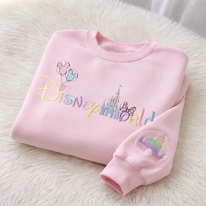 May include: Pink sweatshirt with embroidered "Disney World" text and graphics. Features a castle, balloons, and a bow. The sleeve has a rainbow castle design. The sweatshirt is folded on a white, fluffy surface.