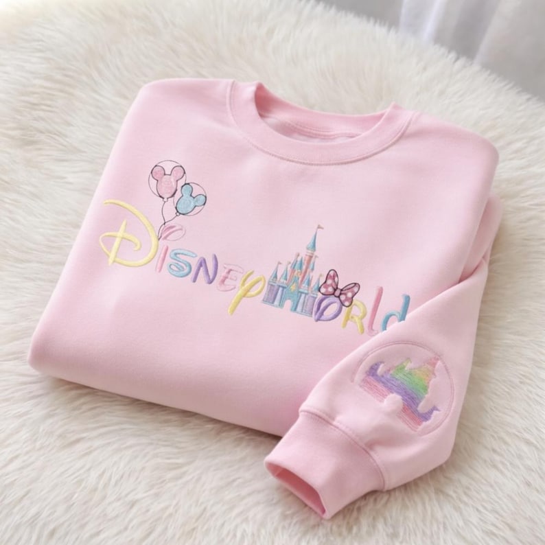 May include: Pink sweatshirt with embroidered "Disney World" text and graphics. Features a castle, balloons, and a bow. The sleeve has a rainbow castle design. The sweatshirt is folded on a white, fluffy surface.