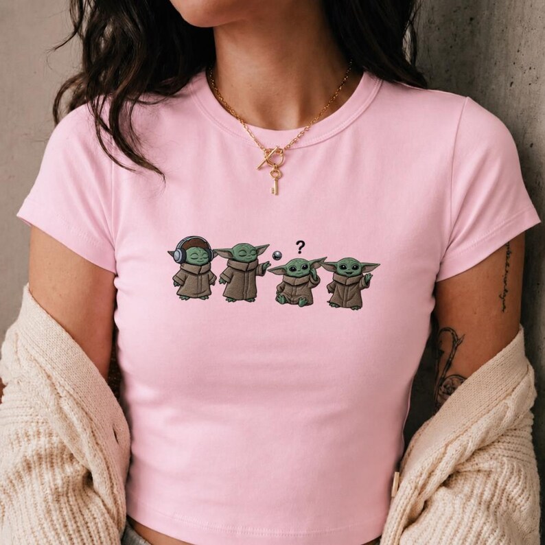 May include: Pink crop top featuring a cartoon graphic of four Grogu characters in various poses. The first Grogu wears headphones, the second and fourth have their arms out, and the third has a question mark above its head.