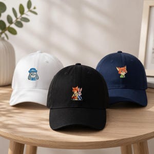 Peut inclure: Trois casquettes de baseball blanches, noires et bleu marine. Chaque casquette présente un personnage de dessin animé brodé différent. La casquette blanche a un lapin, la casquette noire a un renard et un lapin, et la casquette marine a un renard.