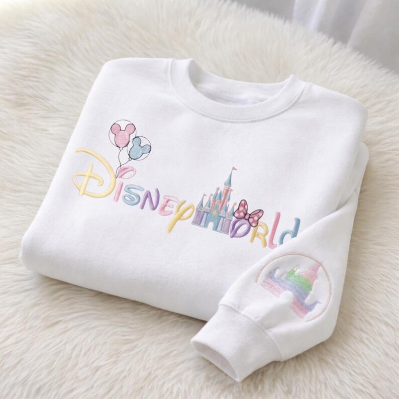 May include: White sweatshirt with embroidered "Disney World" text in pastel colors, featuring balloons, a castle, and a bow. The sleeve has a matching embroidered design. The sweatshirt is folded on a white, fluffy surface.