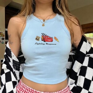 May include: Light blue tank top featuring a graphic of Lightning McQueen from the movie Cars, with Route 66 and checkered flag details. The text "Lightning McQueen" is printed below the graphic. The top has a racerback style.