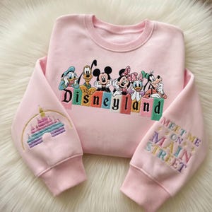 May include: A light pink sweatshirt featuring embroidered Disney characters and the word "Disneyland" in colorful letters. The left sleeve has a castle design, and the right sleeve says "Meet Me on Main Street."