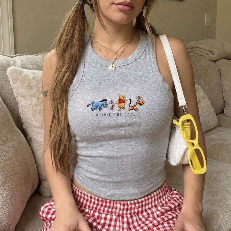Puede incluir: Una camiseta sin mangas gris con un dise&ntilde;o bordado de Winnie the Pooh y sus amigos. El texto "WINNIE THE POOH" est&aacute; debajo de los personajes. La parte superior se combina con una falda a cuadros rojos y blancos.
