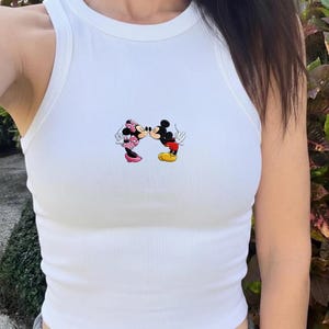 Może przedstawiać: Biały prążkowany top na ramiączkach z kolorową grafiką przedstawiającą całujących się Myszki Miki i Minnie. Postacie z kreskówek mają na sobie swoje klasyczne stroje, Miki czerwone spodenki i żółte buty, a Minnie różową sukienkę w kropki i kokardę.