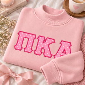 Op de afbeelding: Een lichtroze sweatshirt met een opstaande kraag en grote, geborduurde Griekse letters die "ΠΚΑ" vormen. De letters zijn donkerroze met een wit stippenpatroon en een effen roze rand, gecentreerd op de borst van het kledingstuk.