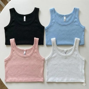 Puede incluir: Cuatro camisetas sin mangas acanaladas en negro, azul claro, rosa y blanco est&aacute;n dispuestas sobre una superficie blanca. Cada camiseta sin mangas tiene un cuello redondo y una peque&ntilde;a etiqueta cerca del escote. Las camisetas probablemente est&aacute;n hechas de un material suave y el&aacute;stico.