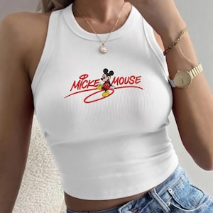 Puede incluir: Camiseta sin mangas blanca acanalada con el texto "Mickey Mouse" en escritura roja y una imagen de dibujos animados de Mickey Mouse. La camiseta sin mangas tiene un cuello redondo y un diseño sin mangas. La parte superior se combina con unos vaqueros azules.