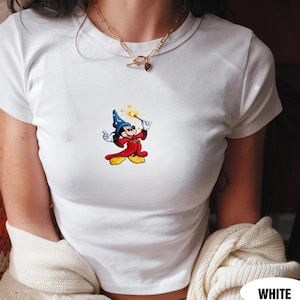 May include: White t-shirt featuring a colorful graphic of a cartoon mouse dressed as a wizard, holding a wand with a starburst effect. The mouse is wearing a blue hat and red robe. The shirt is short-sleeved and has a crew neck.
