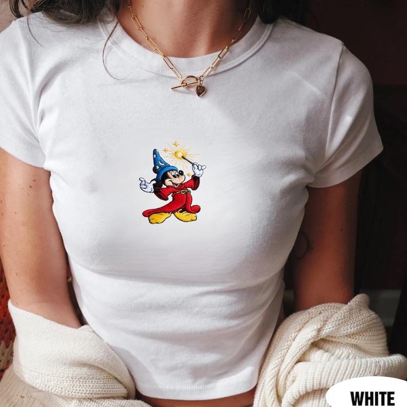 May include: White t-shirt featuring a colorful graphic of a cartoon mouse dressed as a wizard, holding a wand with a starburst effect. The mouse is wearing a blue hat and red robe. The shirt is short-sleeved and has a crew neck.