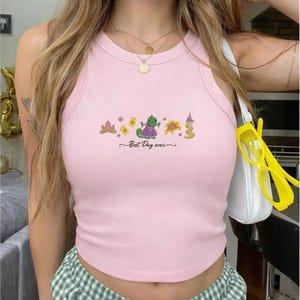 Op de afbeelding: Een lichtroze geribbelde tanktop met een grafische print van een groene kameleon, een bloem, een zon en een toren. De tekst "Best Day ever" is in zwarte cursieve letters onder de kleurrijke illustraties op het midden van de borst gedrukt.