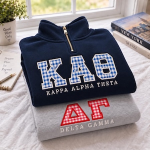 Puede incluir: Un suéter de forro polar azul marino con cremallera de un cuarto, con letras griegas de cuadros azules y blancos para Kappa Alpha Theta, sobre una sudadera gris con letras griegas de cuadros rojos y blancos para Delta Gamma, con texto bordado.
