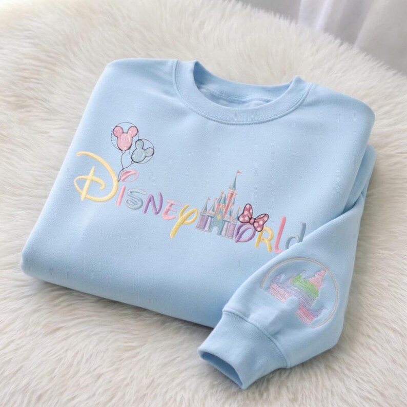 May include: A light blue sweatshirt with embroidered "Disney World" text and graphics. The design includes a castle, balloons, and a bow. The sleeve features a circular design with a castle and rainbow.