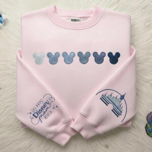May include: A light pink sweatshirt with embroidered Mickey Mouse heads in shades of blue across the chest. The left sleeve features the text "in my Disney mom era" and the right sleeve has a castle design, both in blue embroidery.
