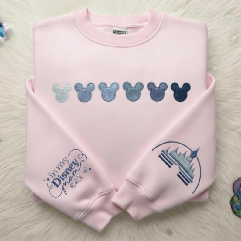 May include: A light pink sweatshirt with embroidered Mickey Mouse heads in shades of blue across the chest. The left sleeve features the text "in my Disney mom era" and the right sleeve has a castle design, both in blue embroidery.