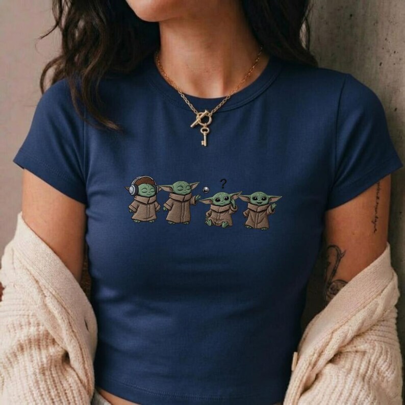 May include: Navy blue crop top featuring a cartoon design of four Grogu characters in various poses. The design includes Grogu with headphones, Grogu with arms outstretched, Grogu with a question mark, and Grogu with a hand raised.