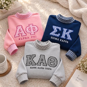 Könnte beinhalten: Drei langärmelige Sweatshirts in Pink, Marineblau und Grau. Jedes hat einen Stehkragen und Bündchen mit Vichy-Karo-Muster. Das pinke Sweatshirt zeigt Alpha Phi, das marineblaue Sigma Kappa und das graue Kappa Alpha Theta.