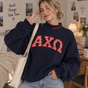 Op de afbeelding: Een marineblauw sweatshirt met lange mouwen en een opstaande kraag met de geborduurde rode tekst Custom Ted met een hartje. De voorzijde toont grote letters AXO in een rood-wit ruitpatroon. Een effen crèmekleurige tote bag wordt gedragen.