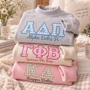 Könnte beinhalten: Ein Stapel aus drei Sweatshirts mit Stehkragen in Grau, Creme und Rosa. Jedes zeigt gestickte griechische Buchstaben mit Vichy-Muster und den entsprechenden Namen der Verbindung darunter: Alpha Delta Pi, Gamma Phi Beta und Kappa Delta.