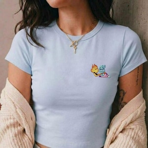 Puede incluir: Camiseta azul claro de cuello redondo con un pequeño gráfico de dos personajes de dibujos animados abrazándose. Los personajes son amarillos y azules con llamas y una cara sonriente. La camiseta es de manga corta.