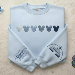 May include: Light blue sweatshirt with embroidered Mickey Mouse heads in varying shades of blue across the chest. The left sleeve features the text "in my Disney mom era" and the right sleeve has a castle design.