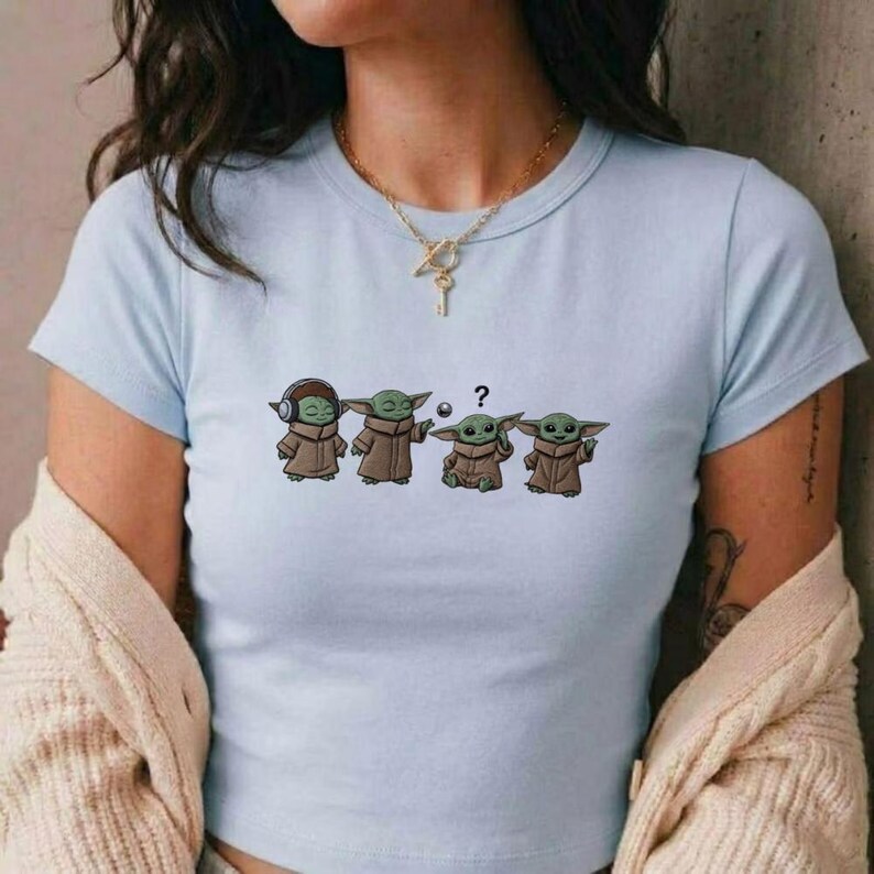 May include: Light blue t-shirt featuring a cartoon design of four Grogu characters in various poses. The first Grogu wears headphones, the second and fourth have their hands up, and the third has a question mark above its head. The shirt is a crew neck.